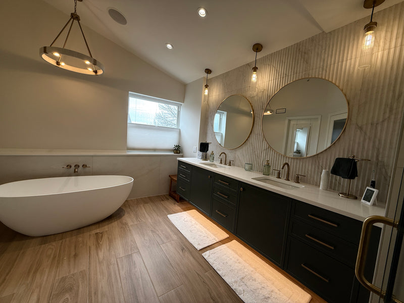 A modern bathroom interior featuring a freestanding white bathtub, a long double-sink vanity with dark cabinets, large round mirrors, wood-look flooring, and warm pendant lighting.